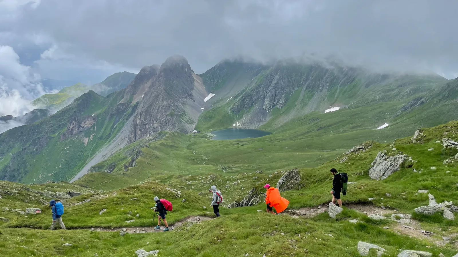 Gruppe von Wanderern in bunten Jacken auf grüner Bergwiese mit Felsgrat und Bergsee unter bewölktem Himmel