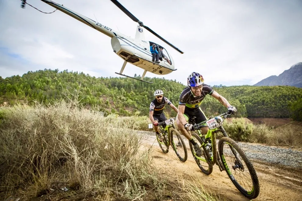 Zwei Mountainbiker fahren auf Sandpiste durch südafrikanische Buschlandschaft, während ein Hubschrauber über ihnen fliegt