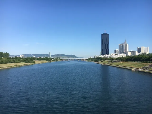 Breiter Fluss mit Grünflächen und moderner Stadtsilhouette unter blauem Himmel an einem sonnigen Tag