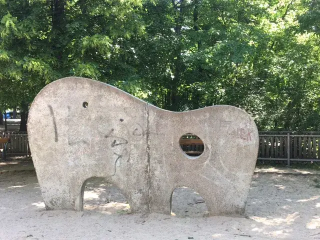 Betonelefant mit Fensteraugen und Rüssel auf Spielplatz, dahinter grüne Bäume und Holzzaun