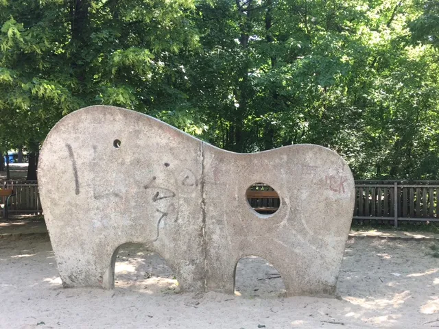 Betonelefant mit Fensteraugen und Rüssel auf Spielplatz, dahinter grüne Bäume und Holzzaun