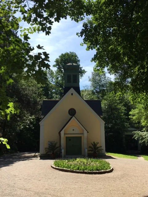Kleine weiße Kapelle mit schwarzem Dach und Schornstein, umgeben von grünen Bäumen und Vegetation, friedliche Atmosphäre