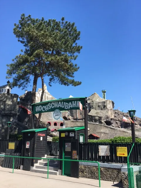 Alte Hochschaubahn im Wurstelprater mit grünem Eingang, markanter Kiefer und Ruine im Hintergrund unter blauem Himmel