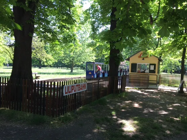 Grüner Praterwald mit Holzzaun, gelber Bahnhofstation und Liliputbahn-Gleisen, Besucher warten auf Abfahrt der Schmalspurbahn